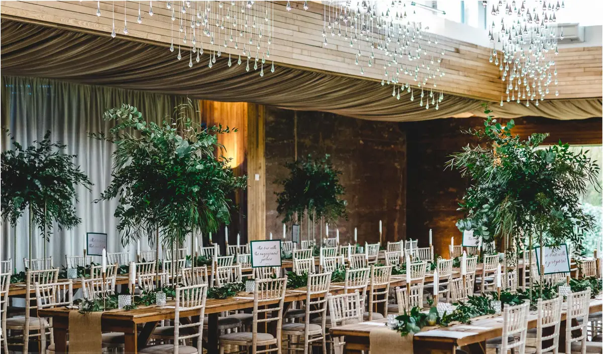 A barn wedding breakfast at Elmore Court: long wooden tables laid with foliage runners and candles, tall greenery arrangements, and strings of hanging lights over a draped ceiling, empty before the guests arrive