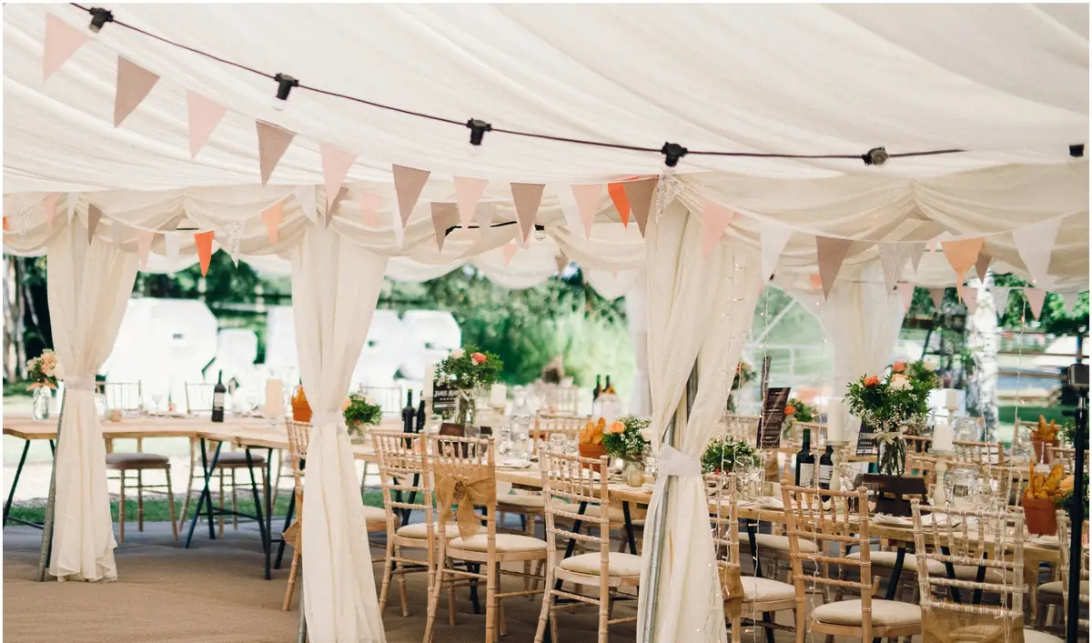 Inside a marquee before the meal: bunting and festoon lights along the roof, tables laid with flowers and wine, and open sides looking on to trees in summer sun